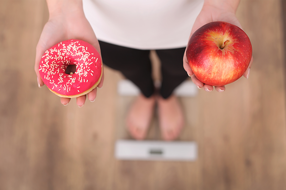 Person on scale holding a donut in one hand and an apple in the other